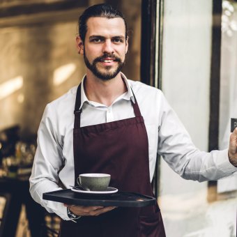 Portrait of handsome bearded barista man small business owner smiling and holding cup of coffee outside the cafe or coffee shop.Male barista standing at cafe